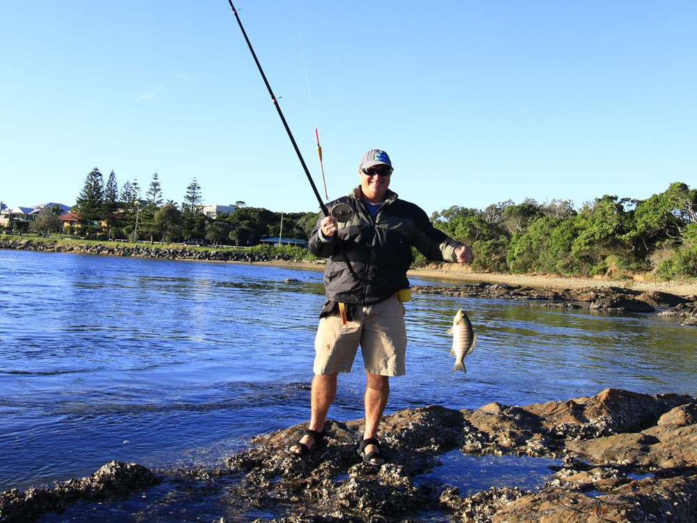 fisherman on rocks holding up a fish that he has caught